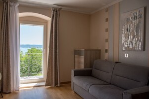 Un salón con sofá, televisión y vistas a un balcón con follaje y una masa de agua.