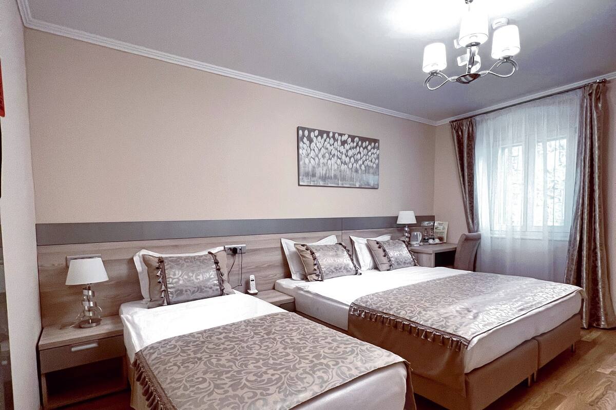 Habitación de hotel con dos camas, una mesita de noche, una lámpara, un cuadro en la pared y una ventana con cortinas.