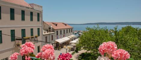 A balcony with pink flowers, a view of a coastal town, and buildings with red roofs.