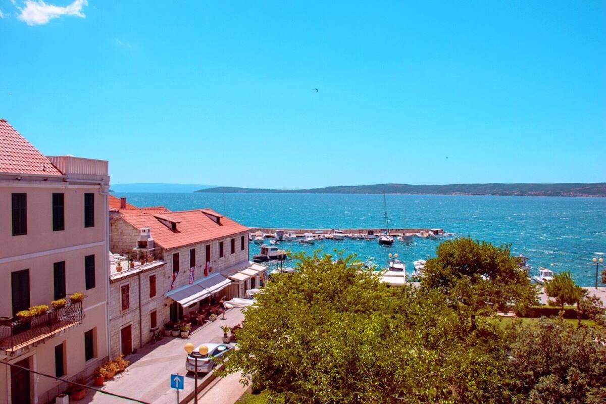 Un pueblo costero con edificios a lo largo de la orilla, un cielo azul despejado y barcos amarrados en un muelle.
