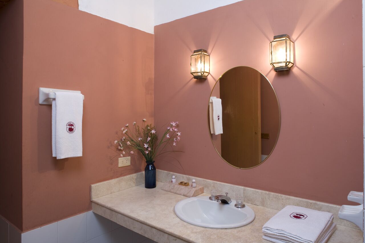 Un baño con encimera color beige, un lavamanos blanco, dos luces fijadas en la pared, un espejo redondo y un florero con flores.