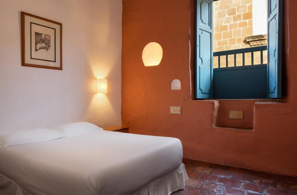 A bedroom with a bed, a wall-mounted light, and a window with blue shutters.