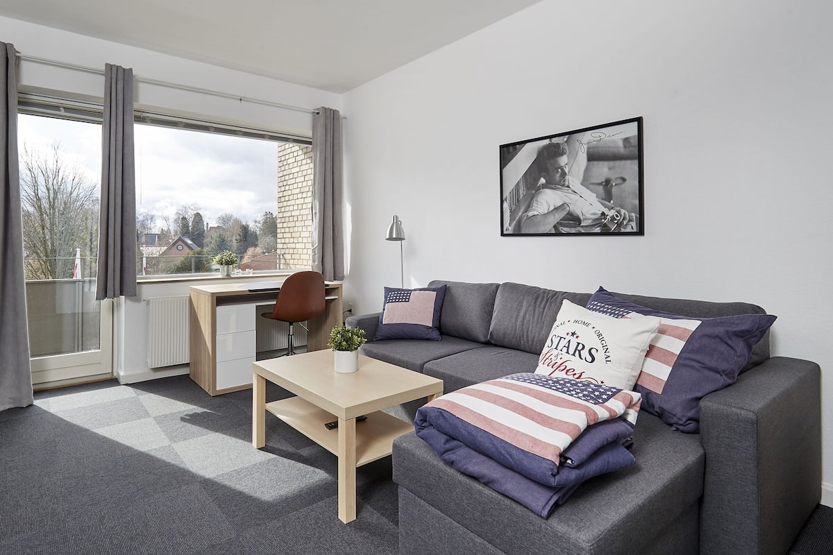 A modern living room with a grey sofa, a small table, a desk, and a large window with curtains.