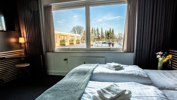 A hotel room with two beds, a window with a view of a building and trees, a radiator, and a lamp.