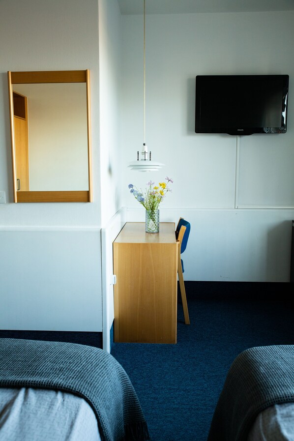 A hotel room with a bed, a desk with a vase of flowers, a chair, a television on the wall, and a mirror.