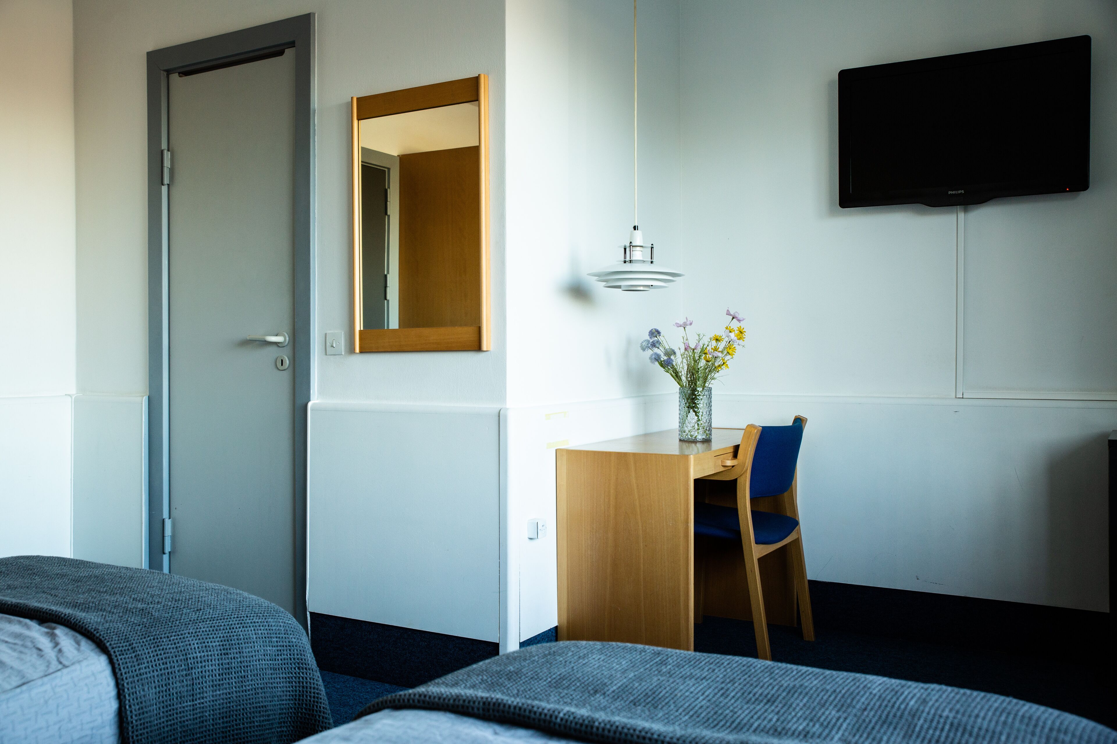 Habitación de hotel con cama, escritorio con silla, espejo, televisor montado en la pared y un florero con flores sobre el escritorio.