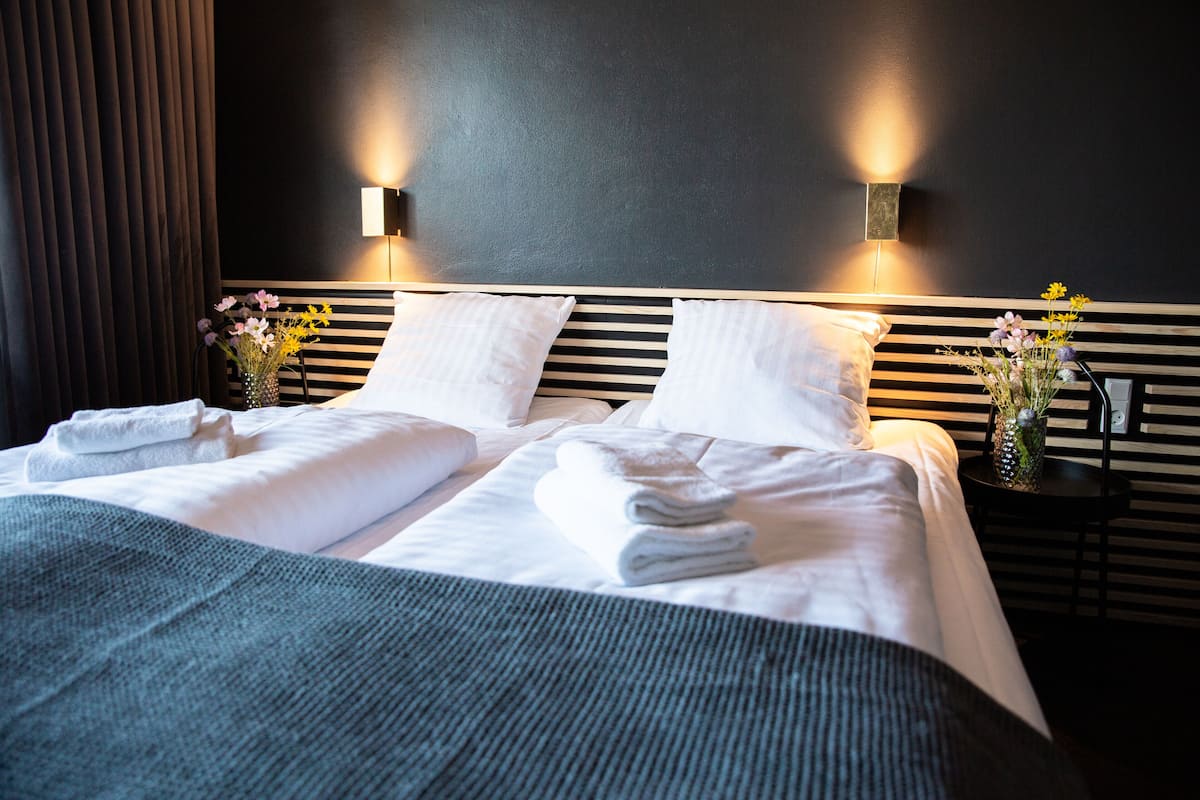A neatly made bed with white linens and towels, two wall-mounted light fixtures, and a vase with flowers on the nightstand.