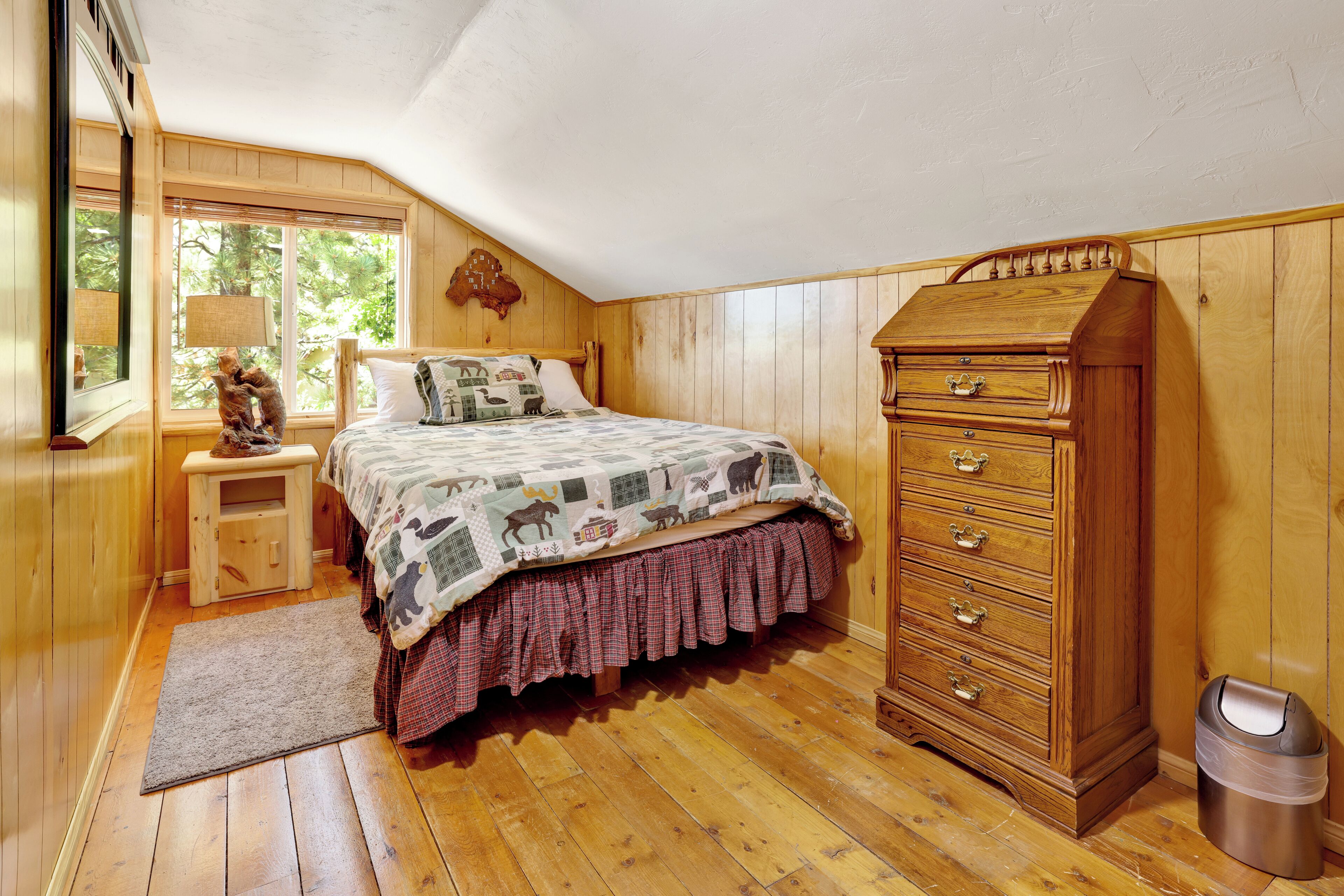 Un dormitorio con piso de madera, una cama con cubrecama estampado, un aparador, una mesita de noche y una ventana con vista a árboles.