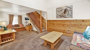 Un salon chaleureux avec un escalier en bois, un tableau accroché au mur et un canapé agrémenté d’un plaid aux couleurs vives.