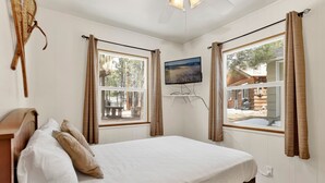 Une chambre à coucher avec un lit, deux fenêtres avec des rideaux, un téléviseur fixé au mur et une tête de lit en bois.