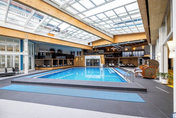 An indoor swimming pool with a glass ceiling, wooden beams, and seating areas.