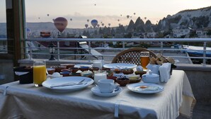 A breakfast table with various dishes and a glass of orange juice, overlooking a scenic view with hot air balloons and unique rock formations.
