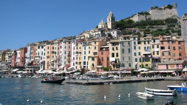 A coastal town with colorful buildings, a historic tower, and a stone fortification on a hill. - La Calata (Portovenere)