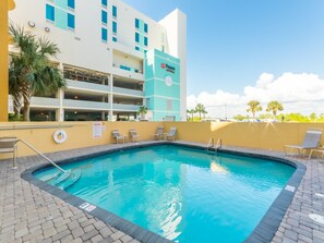 A swimming pool with lounge chairs, a lifeguard stand, and a building with a parking garage in the background. - Emerald Skye 54 (Orange Beach)