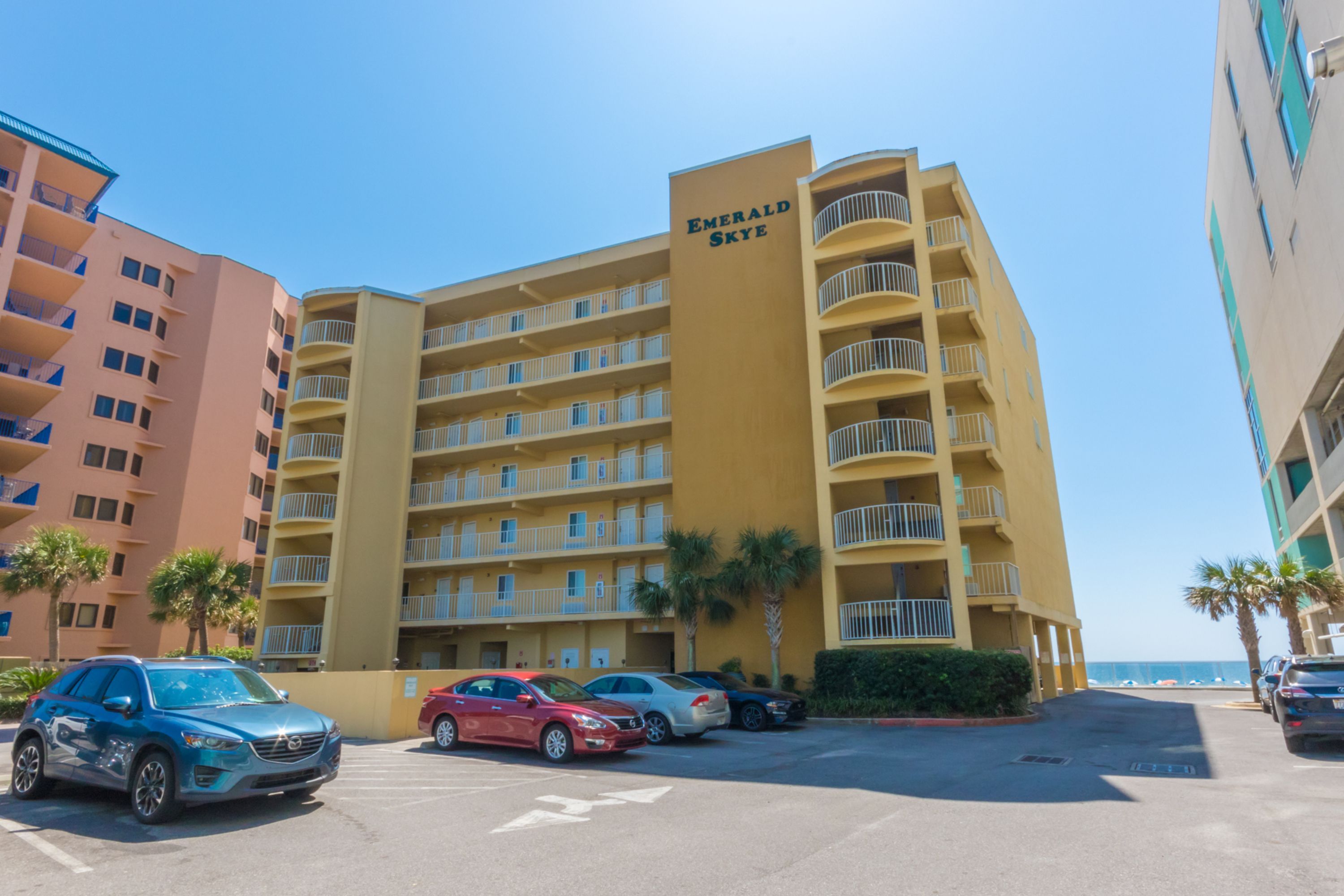 A multi-story hotel with a yellow facade, named 'Emerald Skye,' featuring a parking lot with several cars and a view of the ocean.