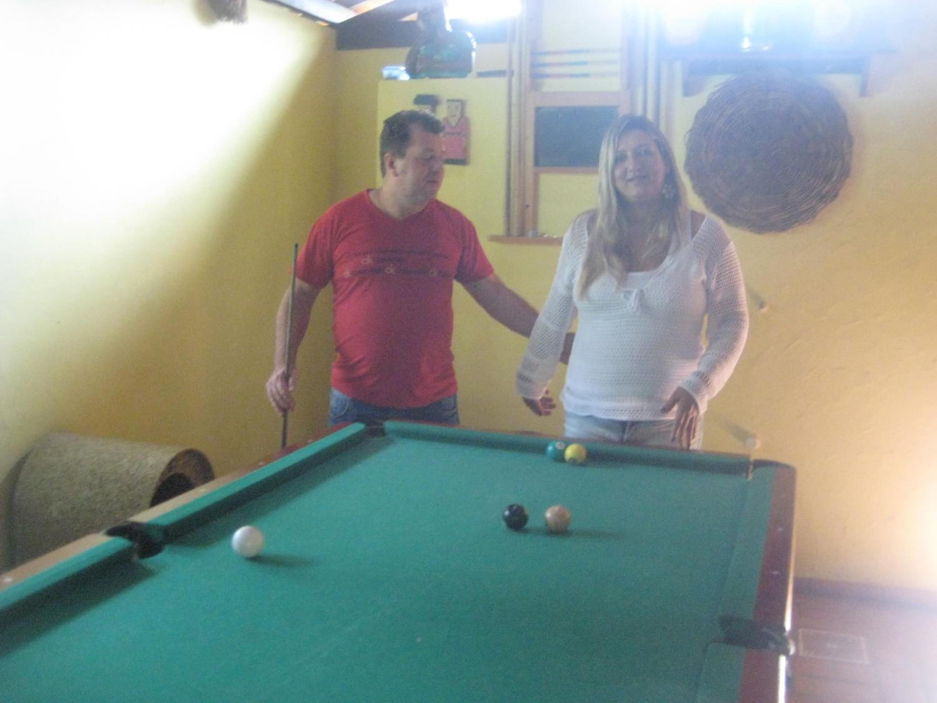 Uma mesa de bilhar com bolas, um homem de camisa vermelha e uma mulher de blusa branca.