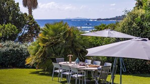 Un espacio para comer al aire libre con mesa y sillas, rodeado de abundante vegetación y vista al mar.