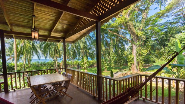 A wooden deck with a table and chairs, surrounded by a railing, overlooking a lush garden and the ocean.