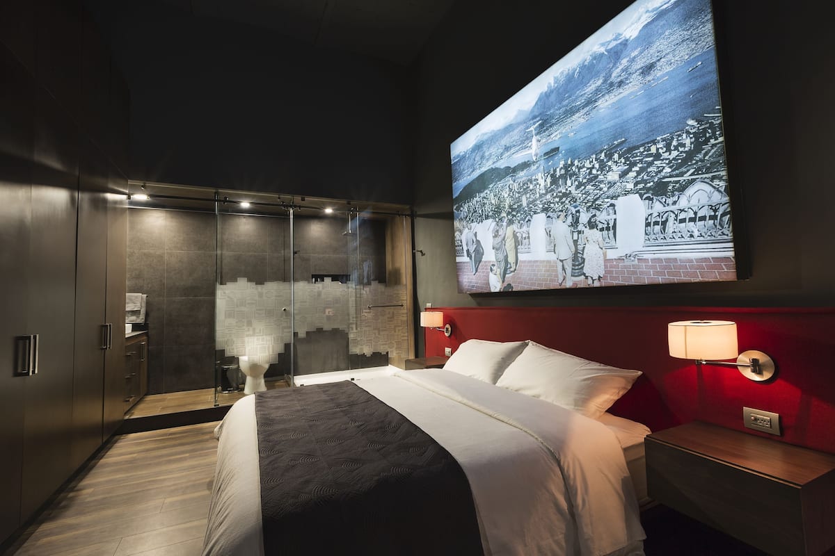 A modern hotel room with a large bed, a glass shower enclosure, a red accent wall, and a cityscape mural.