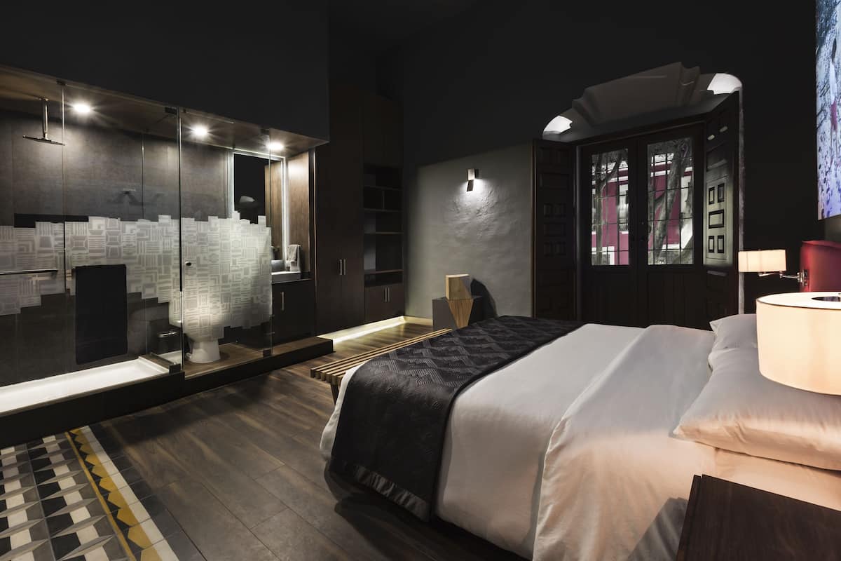 A modern hotel room with a large bed, a glass partition wall, and a view of the cityscape.
