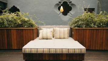 Una cama con estructura de madera y un cojín, flanqueada por dos macetas de madera con plantas verdes.