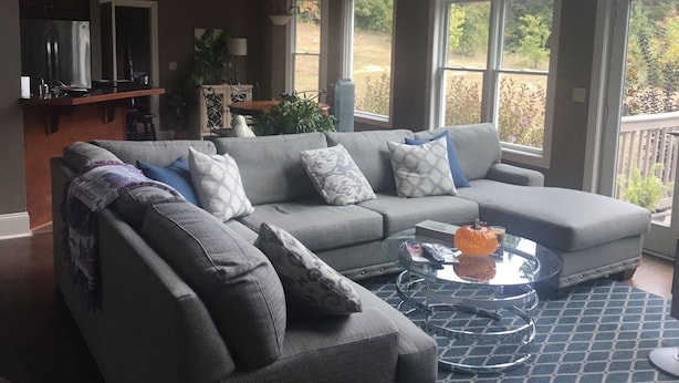 A living room with a grey sectional sofa, a glass coffee table, and a view of the outdoors through large windows. - Beautiful Home close to campus (Oxford)