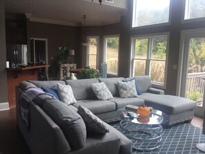 A living room with a grey sectional sofa, a glass coffee table, and a view of the outdoors through large windows. - Beautiful Home close to campus (Oxford)