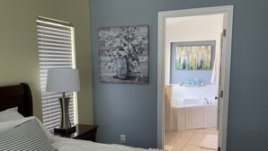Een slaapkamer met een bed, een bedlamp, een schilderij en een zichtbare badkamer door een open deur.