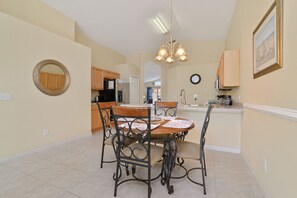 A dining area with a round table, four chairs, and a chandelier. There is a kitchen in the background with wooden cabinets and a refrigerator. - Family Retreat, 4 Bedroom Villa with Private Pool and Spa Near Disney Parks (Davenport)
