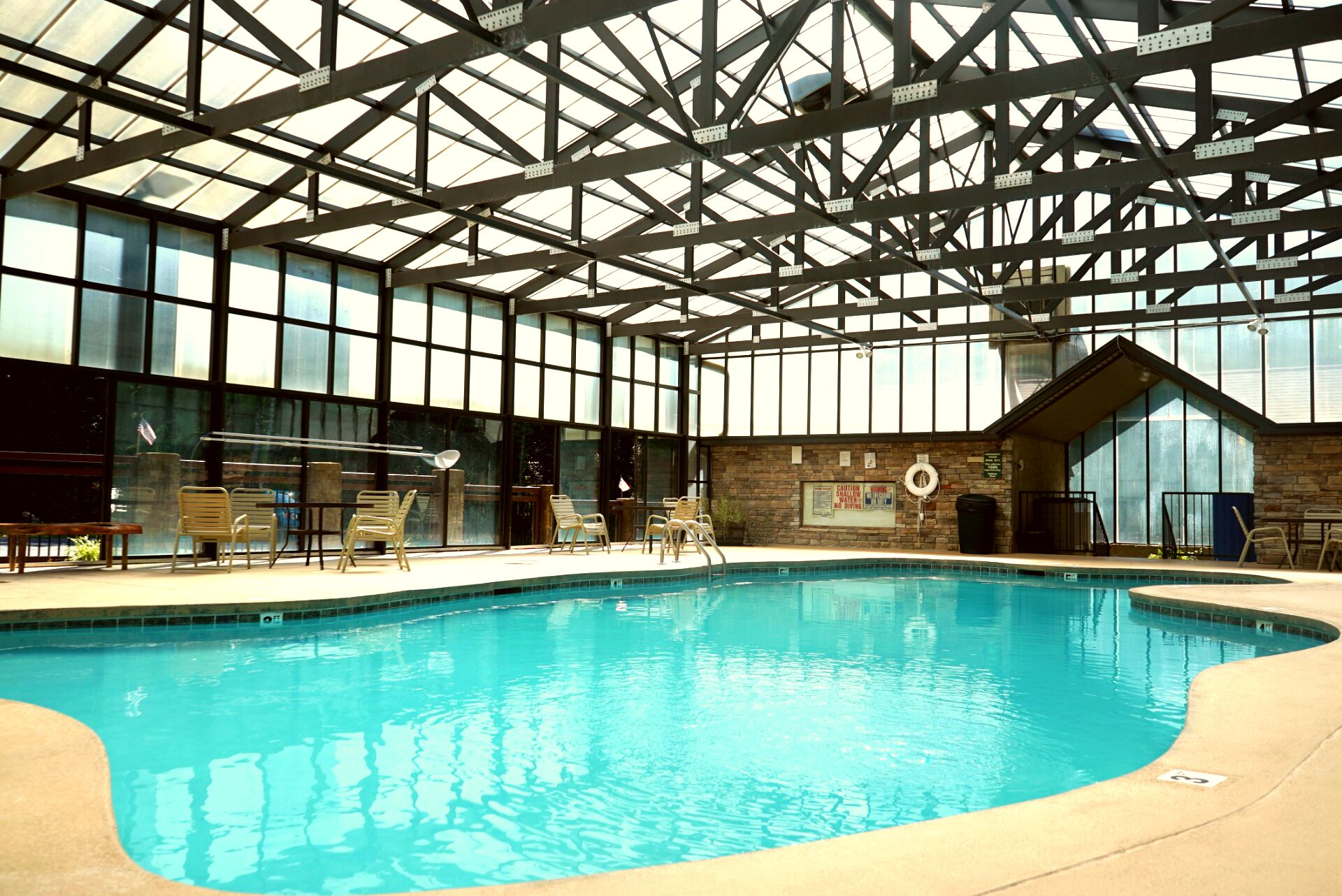 Ein Hallenbad mit hoher Decke und großen Fenstern, umgeben von Liegestühlen und einer Steinwand.