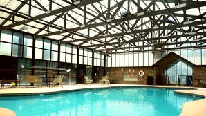 Una piscina interior con techos altos y ventanas amplias, rodeada de sillas de descanso y una pared de piedra.