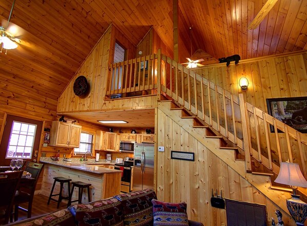 Interior de una cabaña de madera con cocina, comedor y una escalera que sube a un altillo.