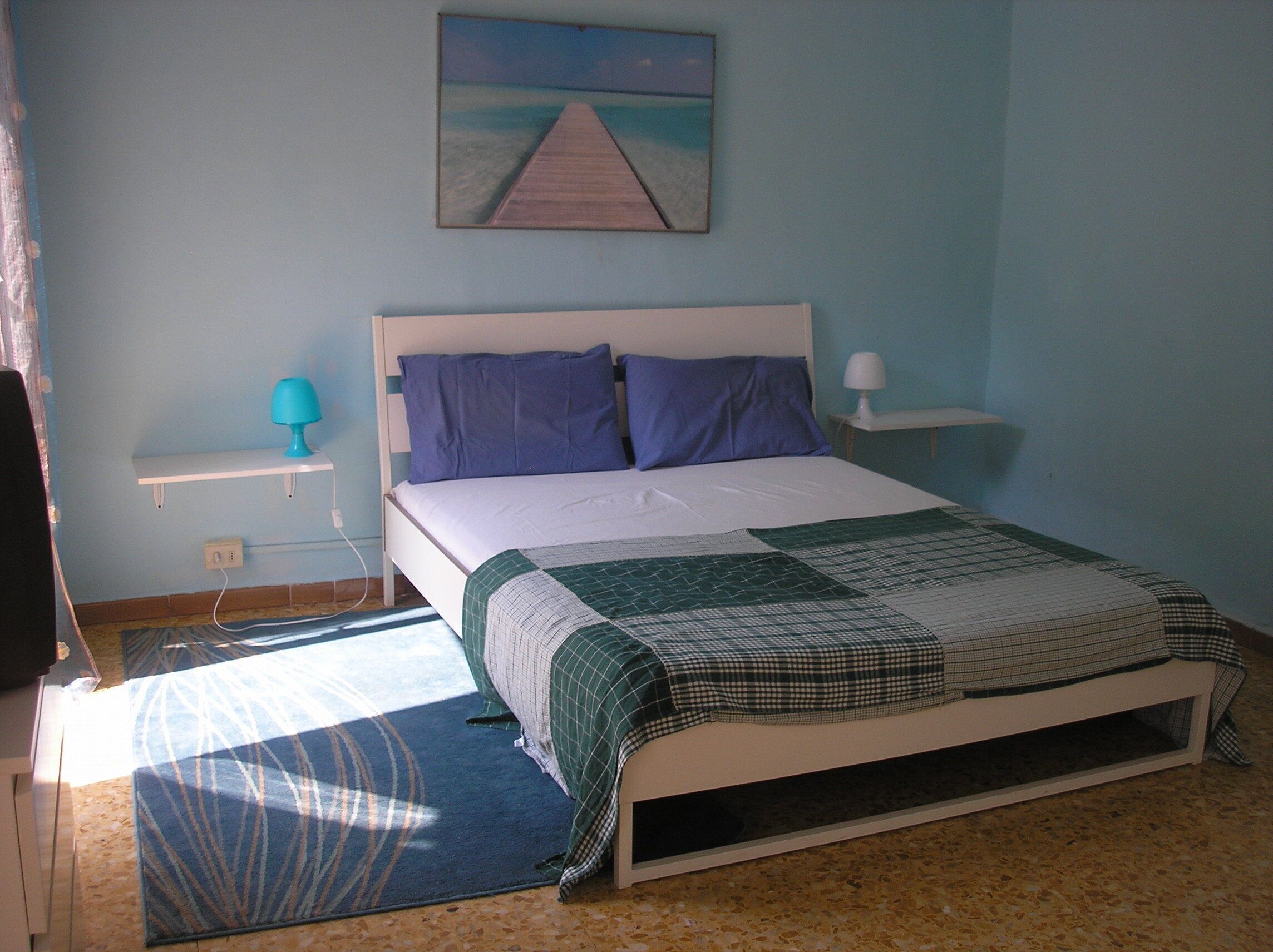 A bedroom with a bed, a nightstand, a lamp, a painting, and a window.