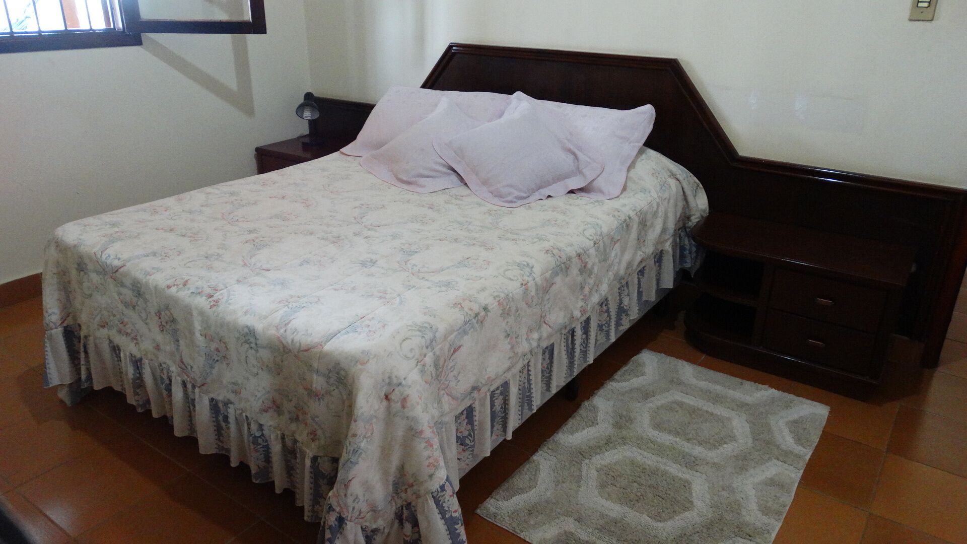 A neatly made bed with a floral bedspread and two pillows, a wooden nightstand with a lamp, and a geometric rug on a tiled floor.