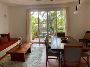 A spacious living area with a sofa, a dining table, and chairs, a coffee table with a flower vase, and a view through a glass door to an outdoor patio with plants.