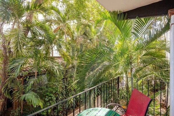 Un balcón con barandilla metálica, rodeado de exuberante follaje verde, incluyendo palmeras y otras plantas tropicales.