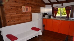 Interior acollidor d'una cabana amb una paret de fusta, un sofà blanc amb coixins vermells, una zona de cuina amb armaris taronja i una finestra amb cortines.