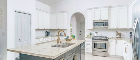 Una cocina moderna con armarios blancos, una isla central con pileta y electrodomésticos de acero inoxidable, además de un revestimiento de azulejos.