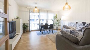 A modern living room with a flat-screen TV, a grey sofa, a dining table with chairs, and two pendant lights. - Urumea | The Rentals Collection (San Sebastián/Donostia)