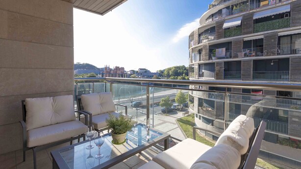 A modern balcony with glass railings, white cushioned seating, and a glass table with a potted plant. - Urumea | The Rentals Collection (San Sebastián/Donostia)