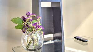 Eine transparente Glasvase mit violetten Blumen auf einem Glastisch vor einer Wand mit einem schwarz-weißen geometrischen Muster.