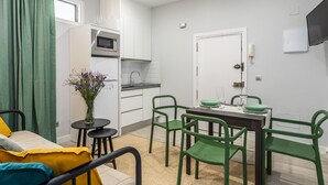 Cocina compacta con armarios blancos, microondas y un pequeño comedor con sillas verdes y una mesa.