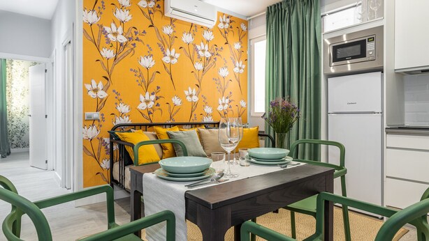 Cocina compacta con microondas, horno y refrigerador. Una mesa de comedor con sillas verdes y un papel tapiz floral amarillo al fondo.