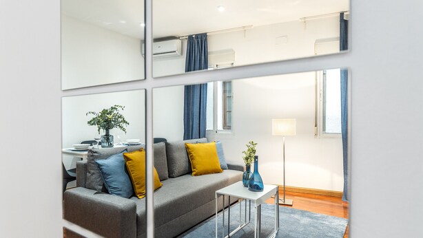 A modern living room with a grey sofa, blue and yellow cushions, a small white table, and a blue rug. - Waou Anton Martin II - Alquiler Corta Duración (Madrid)