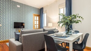 A modern living room with a grey sofa, a dining table set for two, and a large window with blue curtains. - Waou Anton Martin II - Alquiler Corta Duración (Madrid)