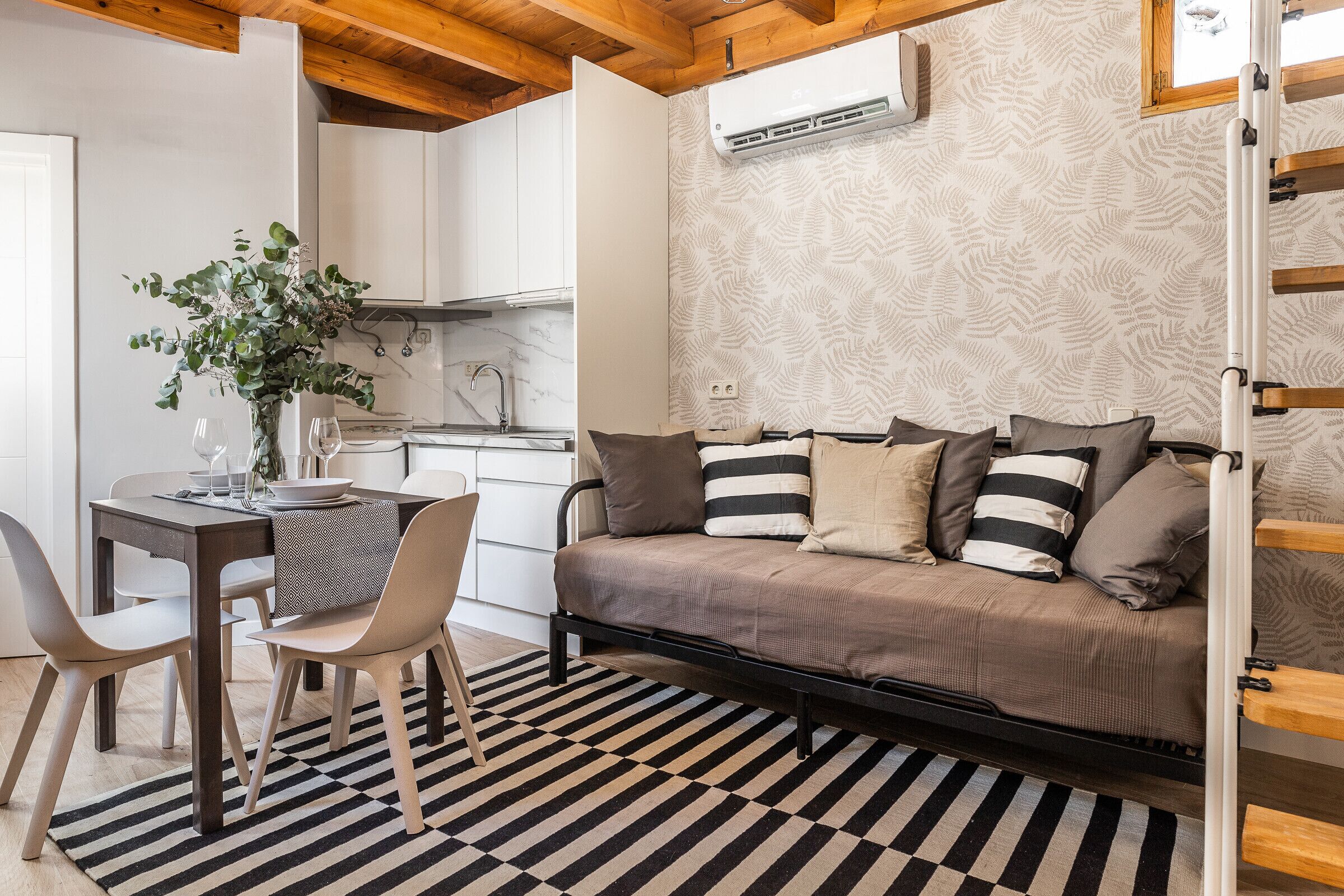 Un espace de vie compact comprenant un canapé, une table à manger et des chaises. Il y a également un coin cuisine avec des meubles blancs et un appareil de climatisation.