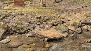 A traditional wooden cabin with a balcony, surrounded by trees and a rocky stream.
