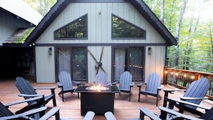 A wooden deck with Adirondack chairs and a fire table, surrounded by a cabin with large windows and a deck railing.