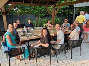 Un grupo de personas cenando al aire libre en una mesa de madera bajo una pérgola.