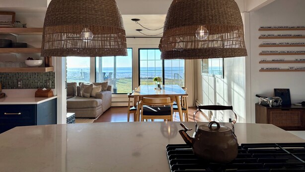 Una cocina moderna con una isla grande, dos lámparas colgantes de mimbre, vista al océano y un comedor con mesa y sillas.